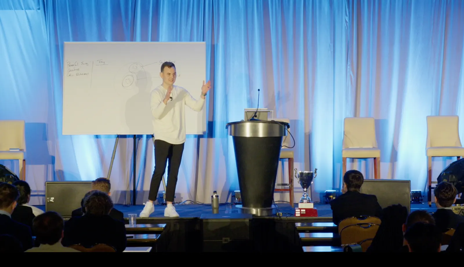 Cory Clark speaking on stage to a seated audience, gesturing beside a whiteboard covered in hand-drawn notes.