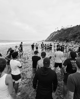 A large group standing in a circle on a beach, listening to someone in the middle of it
